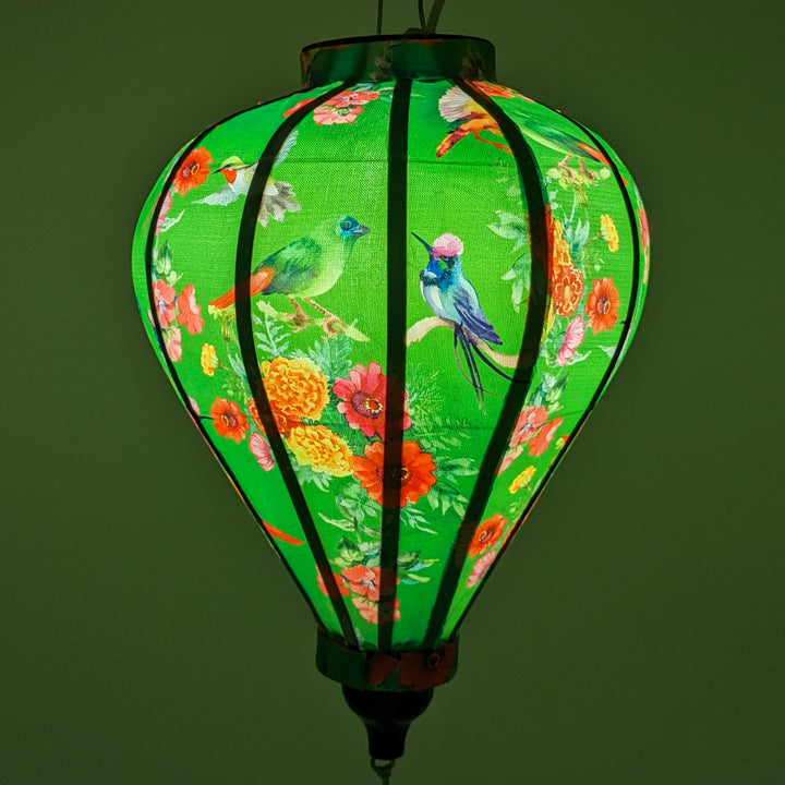 Glowing up close of Balloon shaped lantern with green tassel featuring a print of bright bunches of flowers and various feathery avian species on a bright green backdrop.