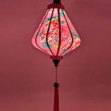 A Teardrop shaped lantern featuring blue birds surrounded by pink and yellow flowers in bloom with a light placed inside creating a soft glowing pink hue.