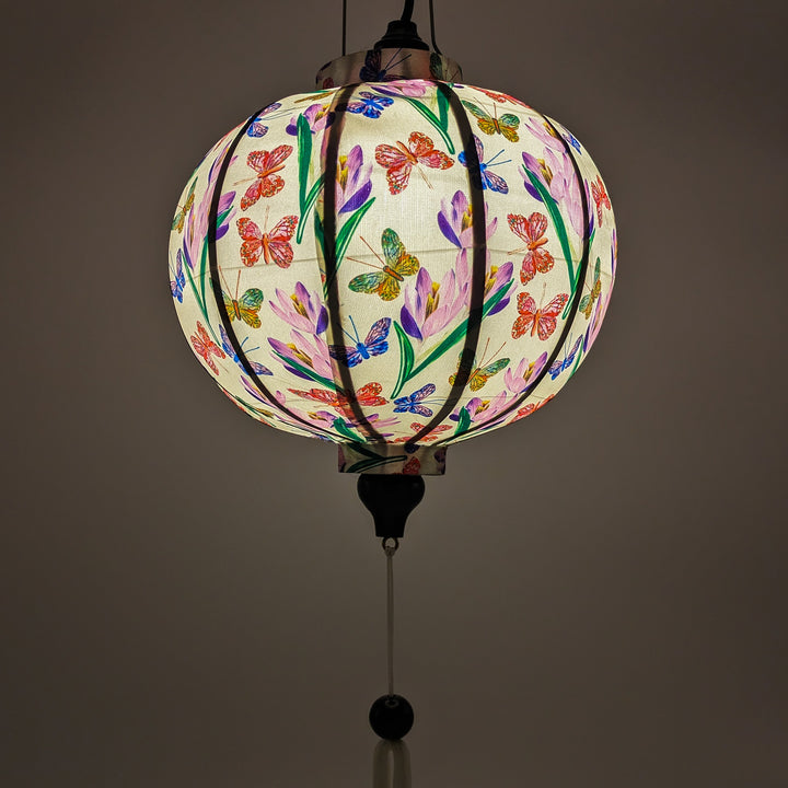 Up close of glowing round shaped lantern with white tassel made with bamboo, steel and a print of stunning print of blue, red and green butterflies fluttering around pink and purple flowers.