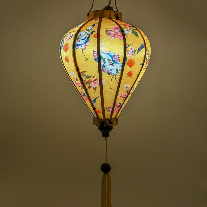 A beautifully illuminated yellow Hoi An silk lantern adorned with intricate blue cranes, delicate pink cherry blossoms, and festive red lantern motifs. The warm glow enhances the elegance of the hand-painted details, while the traditional wooden frame and golden tassel add a classic touch. This lantern brings a cozy ambiance, perfect for home decor, cultural celebrations, or thoughtful gifts.