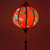 Up close red glowing Round Vietnamese lantern with red tassel with black, yellow and green lanterns and multi colour fans surrounded by white cherry blossoms on a bold red background.