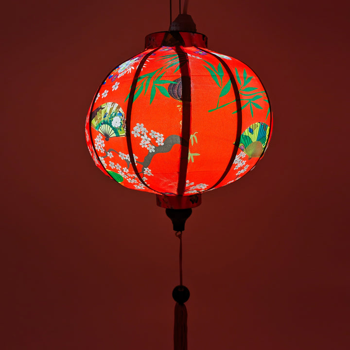 Up close red glowing Round Vietnamese lantern with red tassel with black, yellow and green lanterns and multi colour fans surrounded by white cherry blossoms on a bold red background.