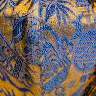 Close up of jacquard silk fabric with intricate patterns of bamboo, lanterns and symbols in blue against gold background.