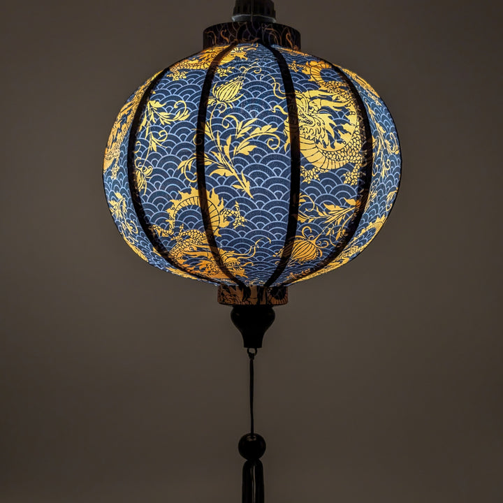 Glowing round Vietnamese lantern with a print of golden yellow dragons and flowers set against a grey sea pattern of swirls on a rich black background.