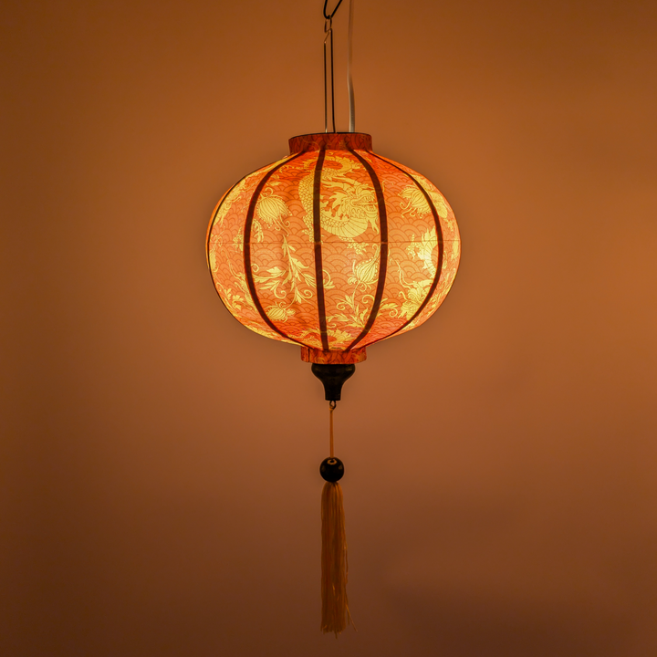 Lit up round Vietnamese silk lantern with golden dragons and orange tassel creating a warm orange glow