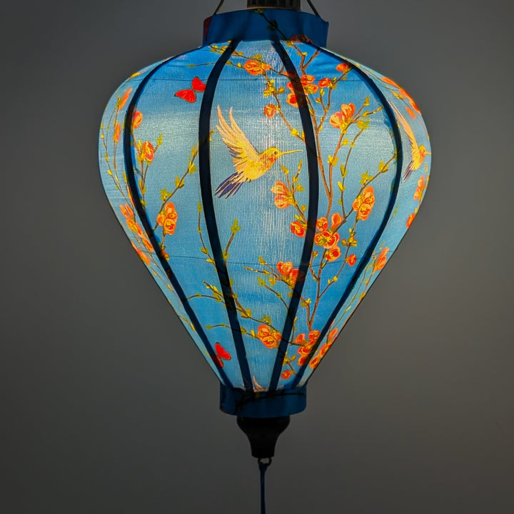 Glowing balloon shape Vietnamese silk lantern featuring a print of hummingbirds in flight with red and pink flowers creating a blue hue.