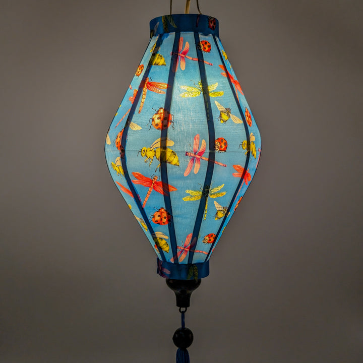 A glowing Tube shaped lantern with a print of dragonflies, lady bugs and bees on bright blue background.