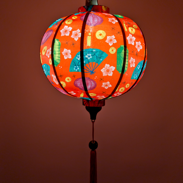 Glowing round silk lantern featuring green lanterns, teal fans, gold coins and pink flowers on red lantern with matching red tassel.