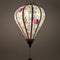 A glowing balloon shaped Vietnamese lantern made in Hoi An with pink and black cranes among lush green foliage and delicate blooming flowers. 