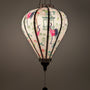 A glowing balloon shaped Vietnamese lantern made in Hoi An with pink and black cranes among lush green foliage and delicate blooming flowers. 
