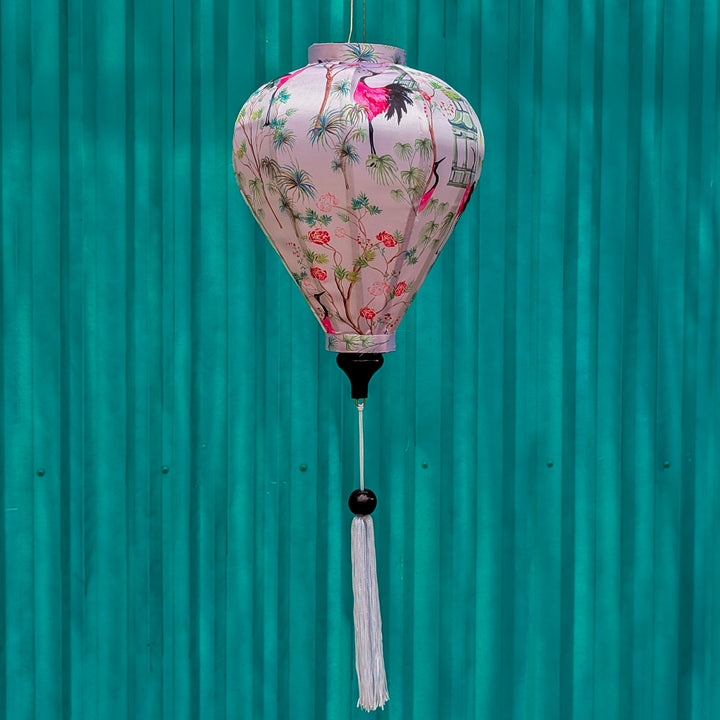 A balloon shaped lantern hanging in front of a bright blue wall featuring pink and black cranes among lush green foliage and delicate blooming flowers. 