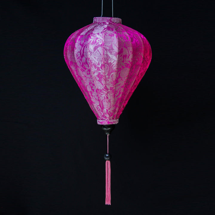 A balloon shaped lantern with pink tassel made from jacquard silk fabric with intricate patterns of bamboo, lanterns and symbols in silver against pink.