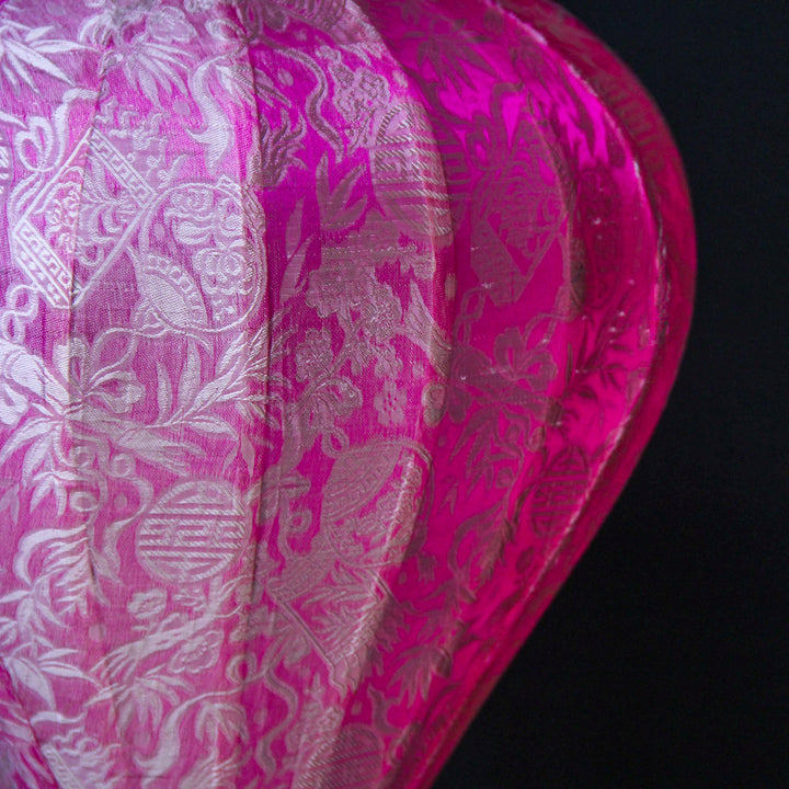 Close up of a lantern made with jacquard silk fabric with intricate patterns of bamboo, lanterns and symbols in silver against pink background.