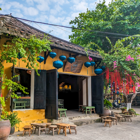 Traditional yellow building with open doors, tables, and chairs in a street setting with greenery and teal silk lanterns and red decorations in the tree.