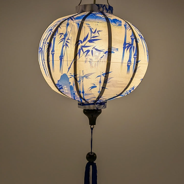 Glowing round Vietnamese silk lantern hand crafted in Hoi An, featuring a blue print of temples and pagodas high above hilltops with large bamboo trees floating against white background. 