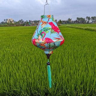 Teardrop shaped lantern with a print of pink flamingos on a light blue background hanging in a rice field.