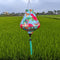 Teardrop shaped lantern with a print of pink flamingos on a light blue background hanging in a rice field.