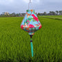 Teardrop shaped lantern with a print of pink flamingos on a light blue background hanging in a rice field.