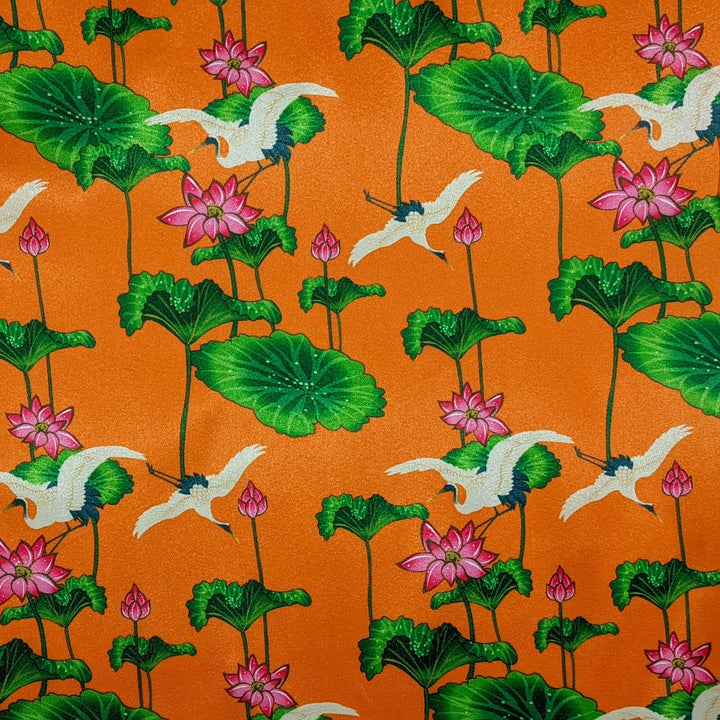 Orange polyester silk fabric with white cranes surrounded by pink flowers with large green leaves.